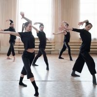 Gruppe von f&uuml;nf T&auml;nzerinnen und T&auml;nzern in schwarzer Kleidung in einem hellen Tanzstudio mit gro&szlig;en Fenstern, die synchron mit erhobenen Armen und ausgestreckten Beinen tanzen