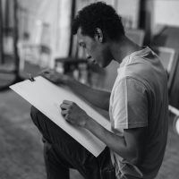Junger Mann mit einem gro&szlig;en Blatt Papier sitzt in einem Atelier und zeichnet