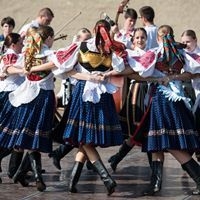 Menschen in traditioneller Tracht tanzen im Kreis mit verschr&auml;nkten Armen auf einem Platz