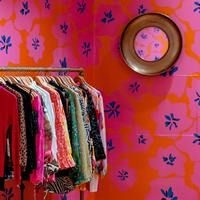 Kleiderstange mit verschiedenen bunten Kleidungsst&uuml;cken vor einer orangefarbenen Wand mit blauem Blumenmuster und rundem Spiegel