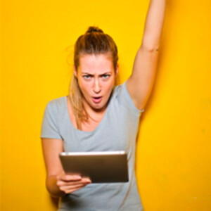 Eine junge Frau steht jubelnd mit ihrem tablet vor einer gelben Wand.